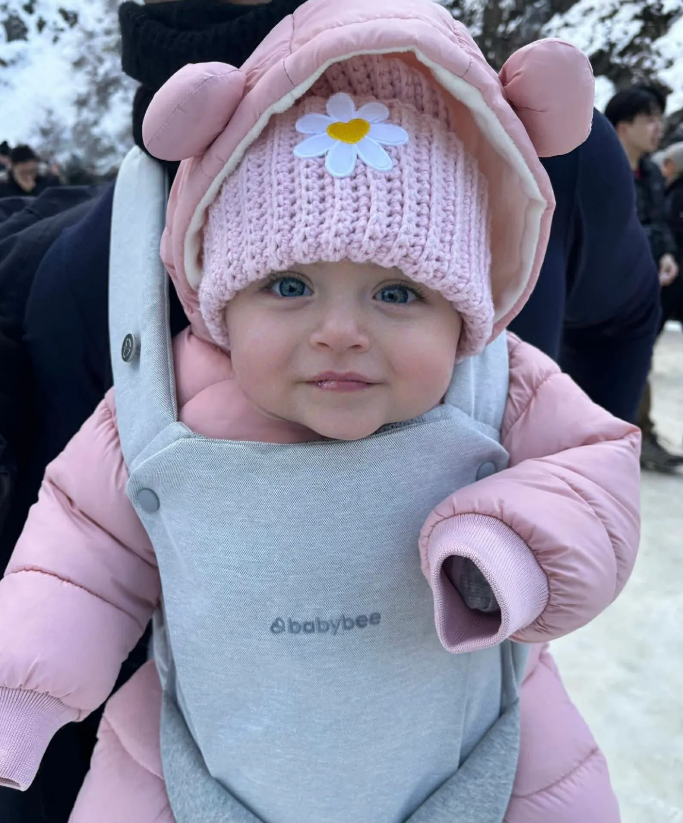 A small child in a pink puffer being worn in a carrier, wearing a soft pink beanie with a white daisy patch.