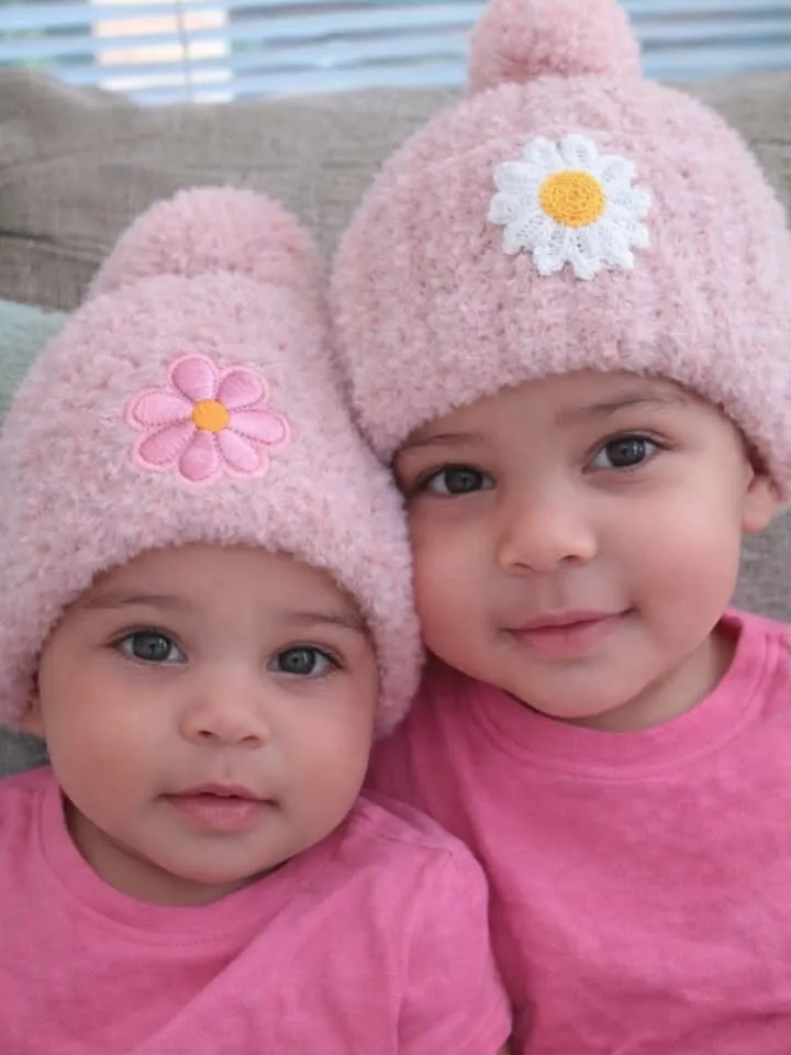 Two small children in pink shirts, each wearing a soft pink beanie with a daisy patch.
