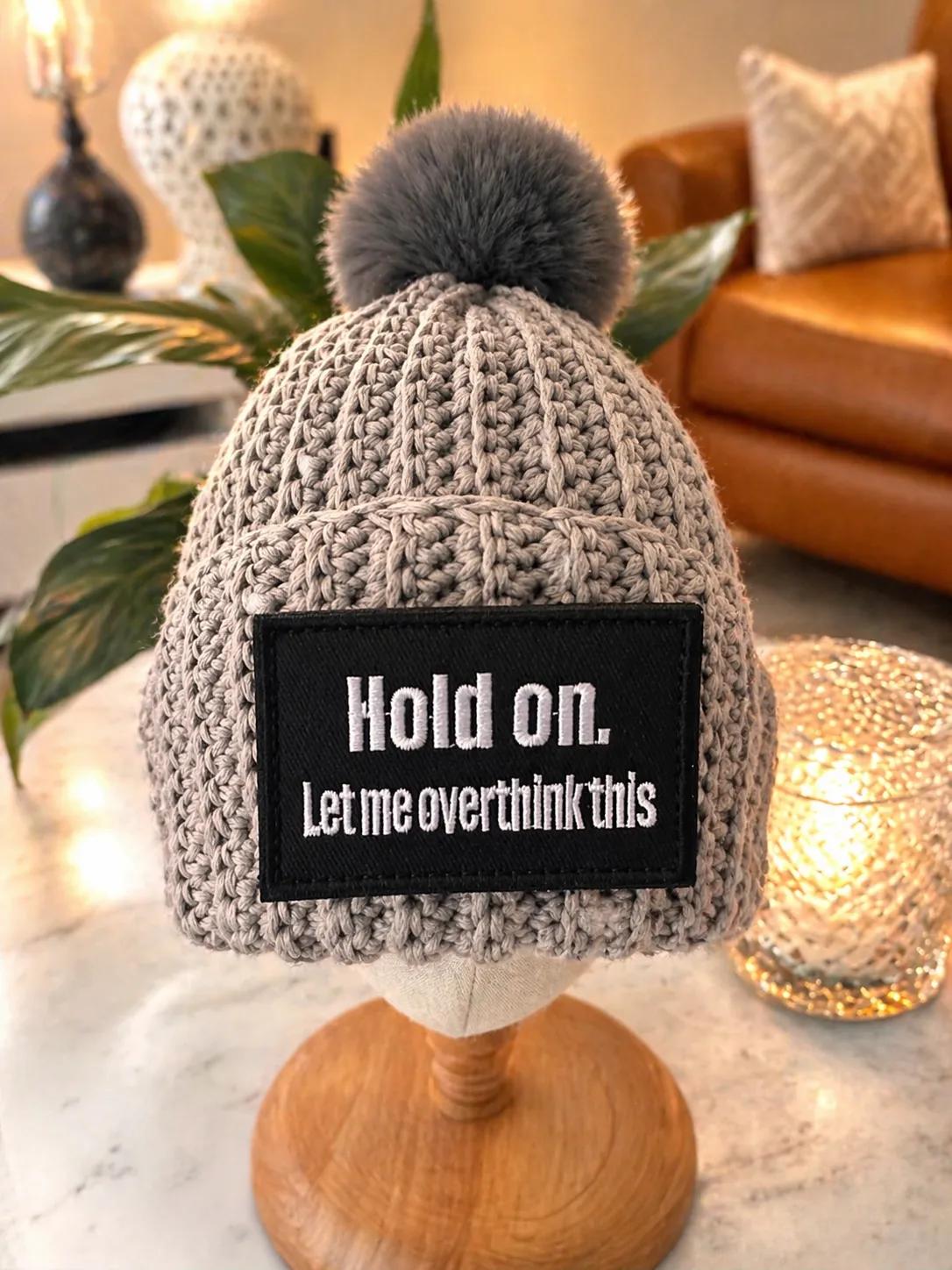 Grey cable-knit beanie with dark grey pompom, displayed on a wooden stand. A black woven patch on the front reads Hold on. Let me overthink this