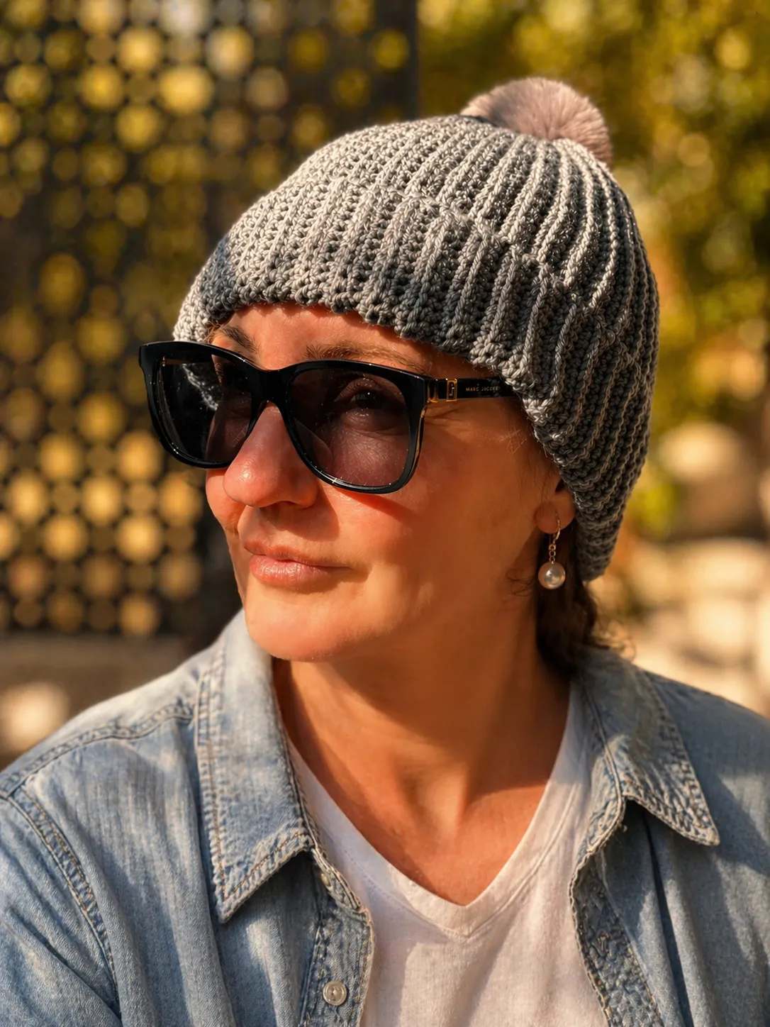 Adult wearing a grey ribbed crochet beanie with a dusty rose pom pom, paired with sunglasses and a denim shirt