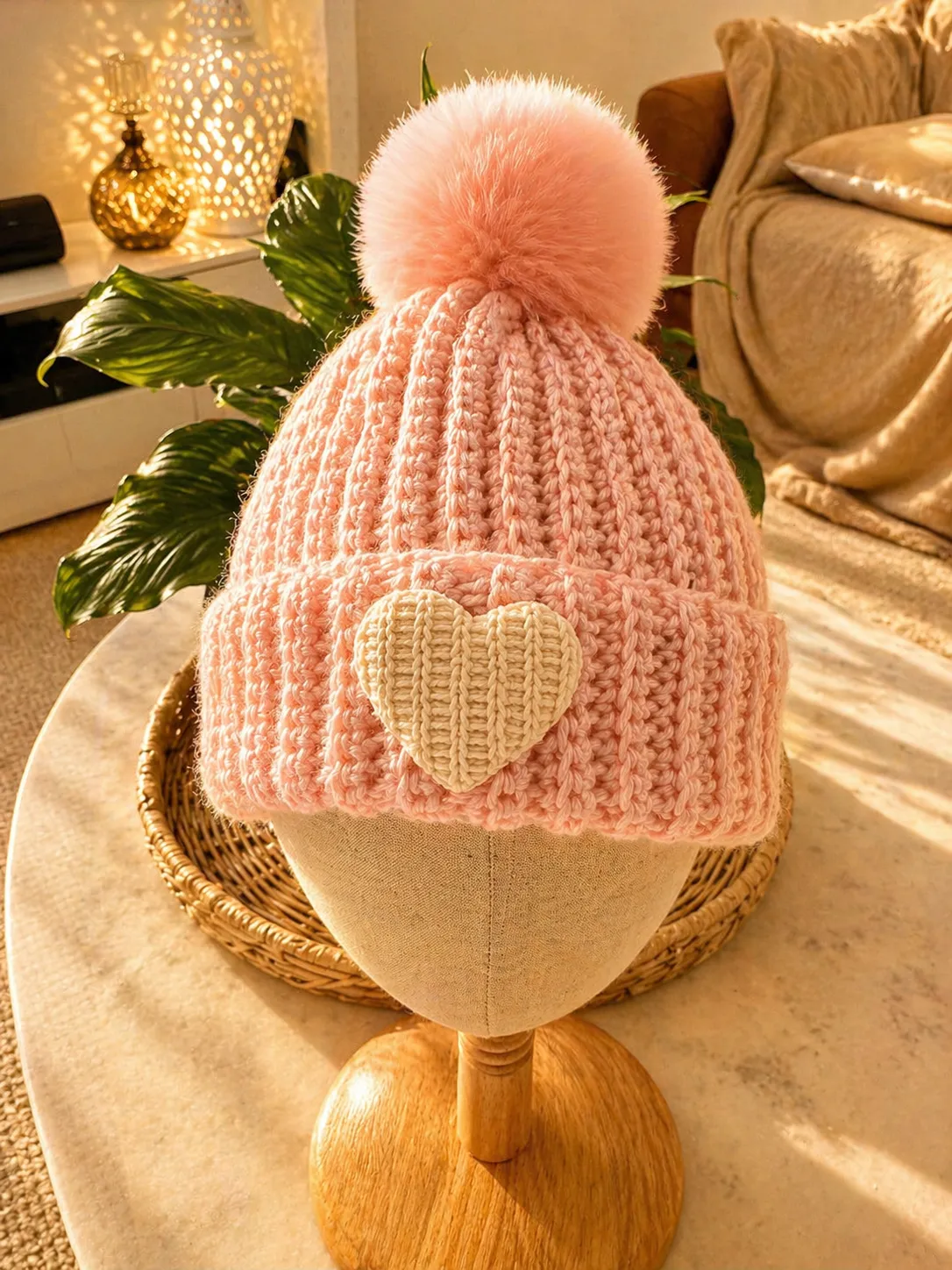 Pink hand-crocheted beanie with ribbed texture, cream-coloured heart patch on the front, and a large fluffy pink pom pom on top, displayed on a wooden stand in a warm home setting