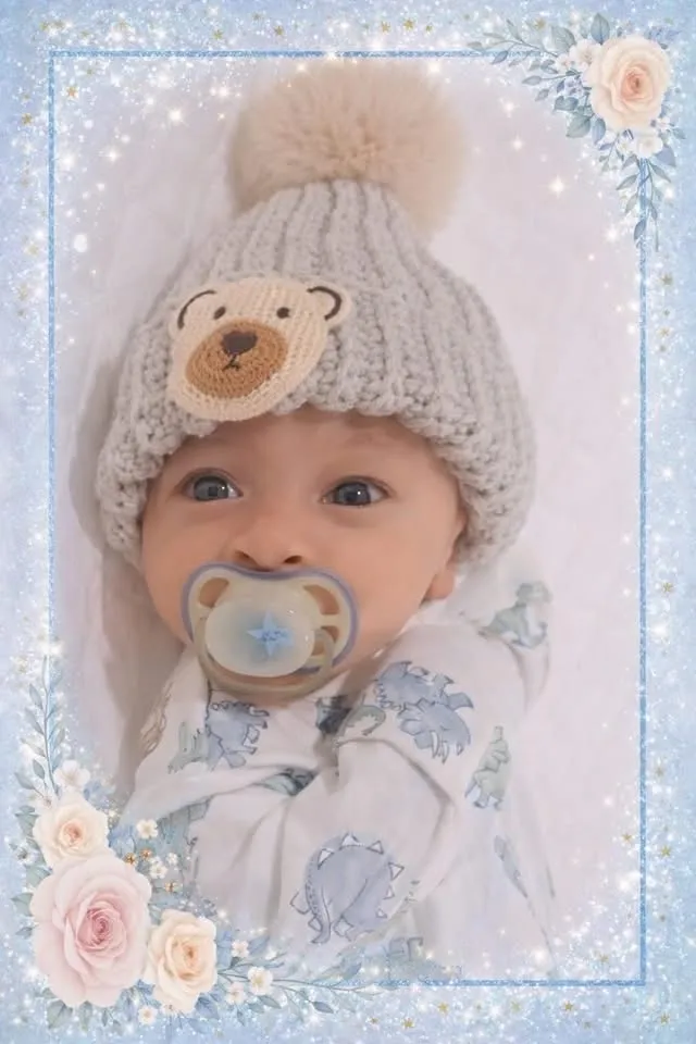 A newborn baby wearing a light-grey ribbed beanie with a crocheted bear face and cream pom.