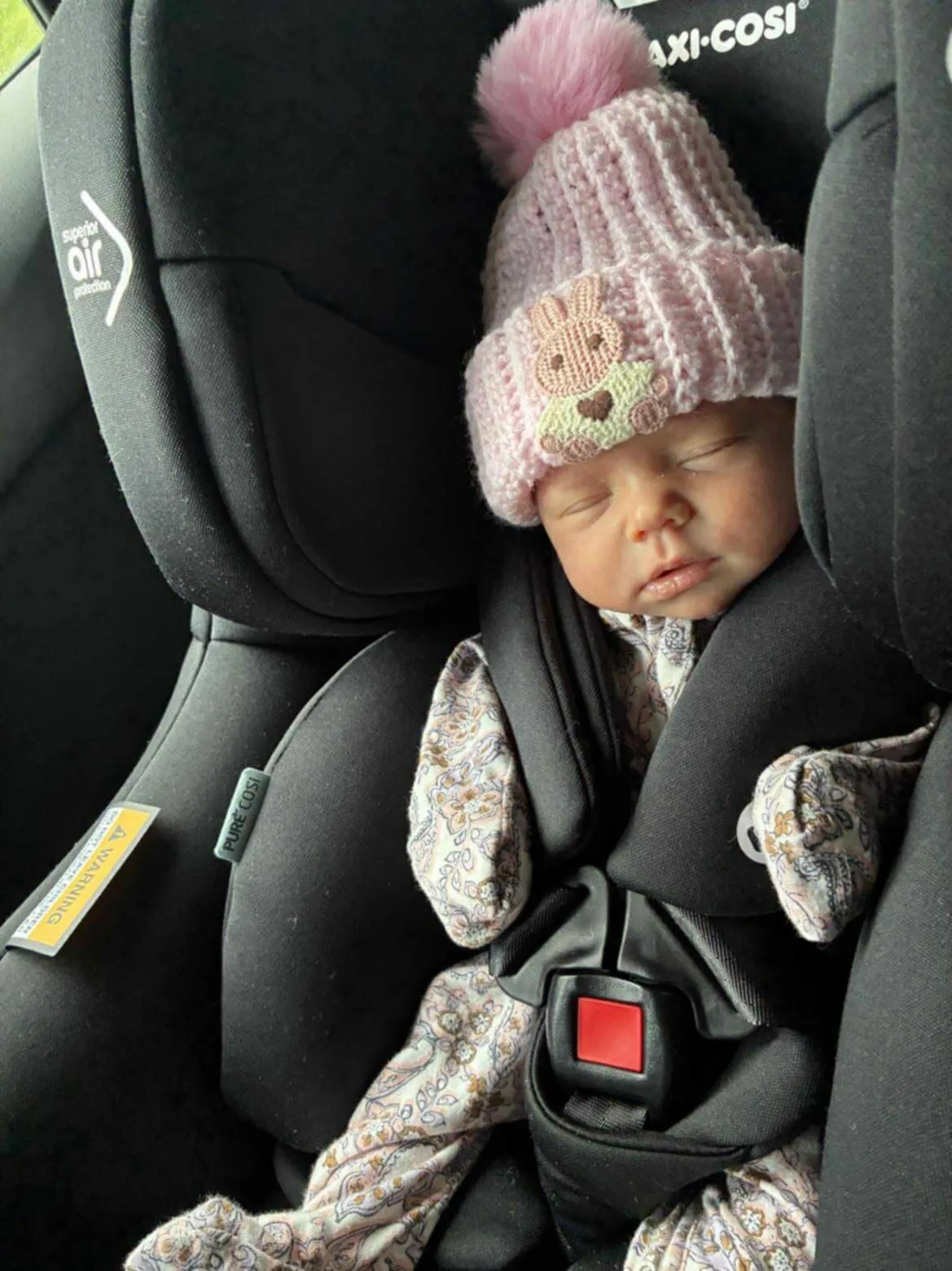 A newborn asleep in a car seat, wearing a soft pink ribbed beanie with a bunny patch and pink pom.