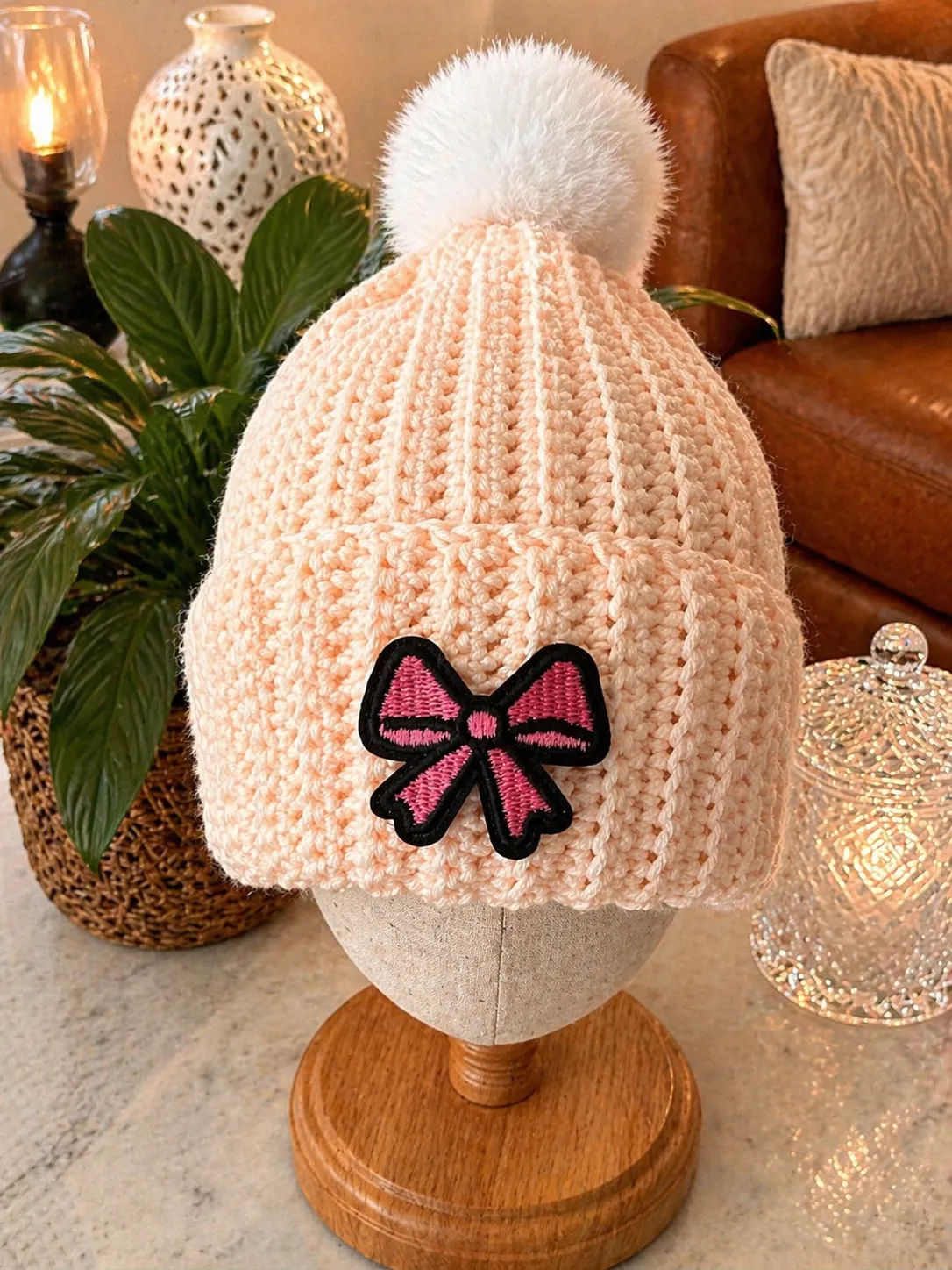 Blush pink crocheted beanie with white pom pom and embroidered pink bow with black outline, displayed on a wooden head form in a styled home setting