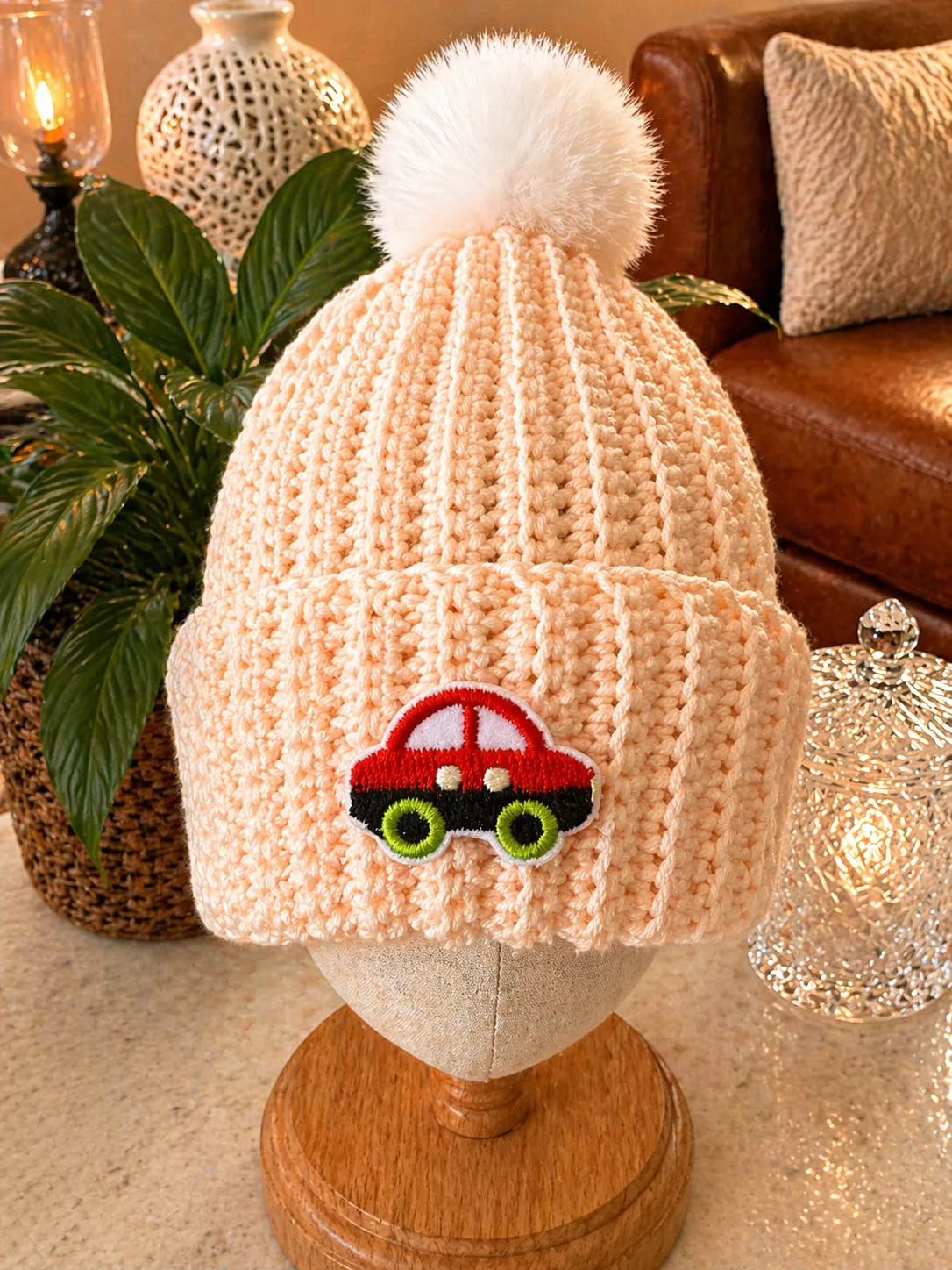Hand-crocheted blush pink ribbed beanie with white pom pom, featuring a red car with black body and lime green eyes embroidered on the front
