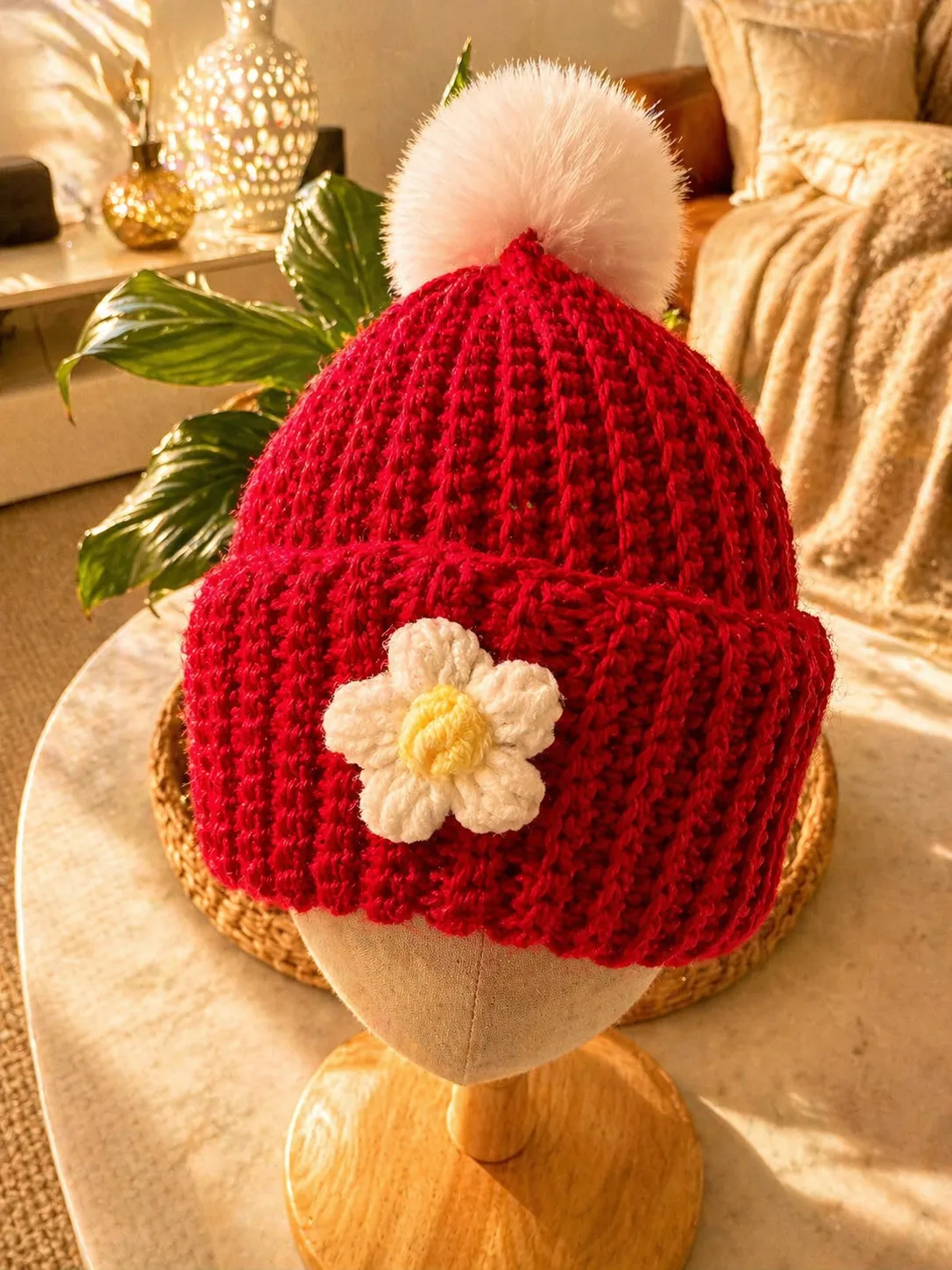 Hand-crocheted red baby beanie with cream pom pom on top and white daisy flower with yellow centre appliqué on the front, displayed on a wooden head form.