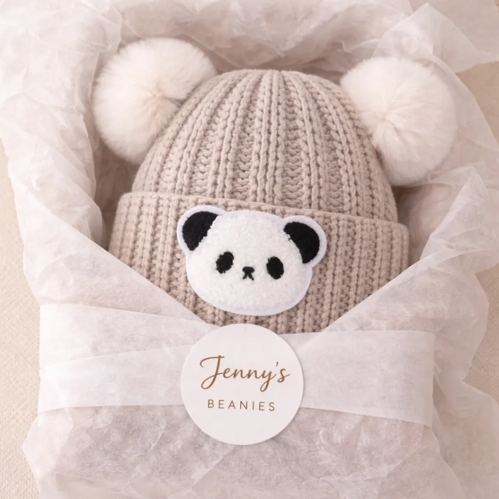 An oatmeal beanie with two white pom ears and a panda face, wrapped in tissue with a circular Jenny's Beanies sticker.